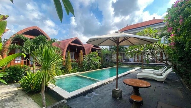 Zona de piscina con una tumbona y una sombrilla, rodeada de exuberante vegetación y un edificio con tejado rojo.