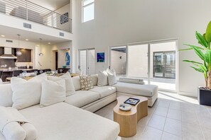 A spacious living area with a large sofa, a coffee table, and a dining table set for four. There is a balcony with a sliding glass door, and a kitchen area is visible in the background.