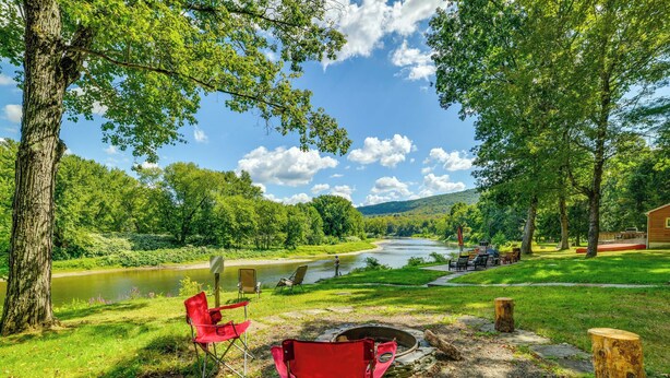 Un camping con sillas rojas, un hogar para fogata y vistas a un río y bosque.