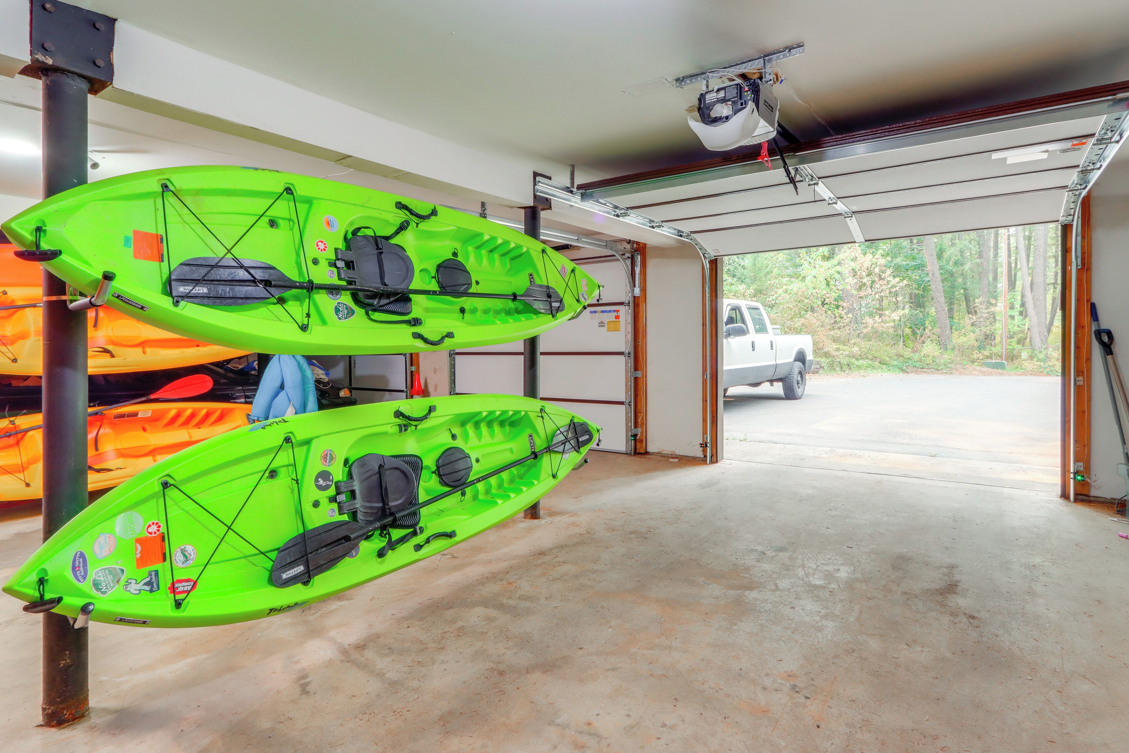 Un garaje con dos kayaks montados en soportes, una camioneta blanca estacionada afuera y vista de árboles a través de la puerta del garaje.
