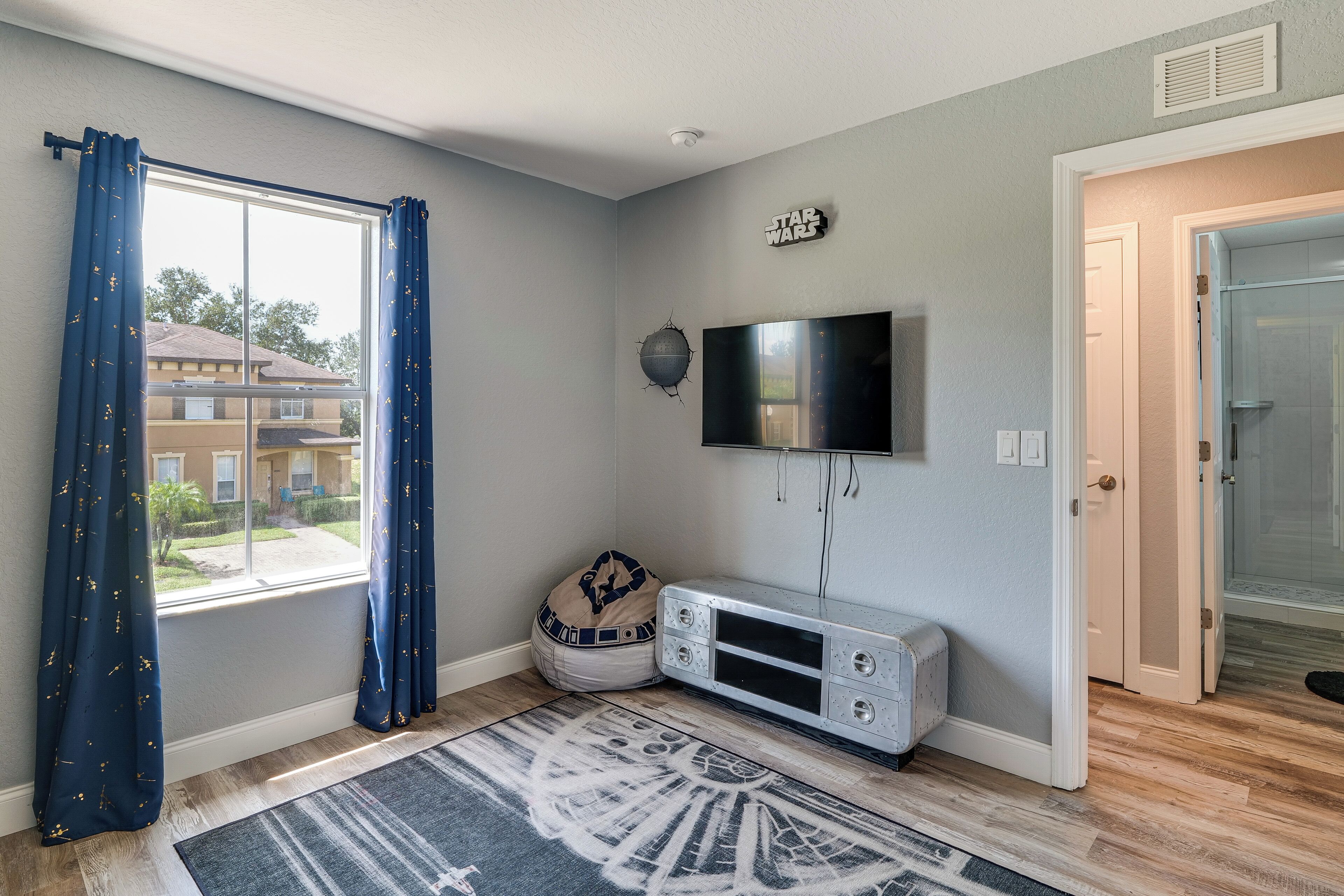 Una habitación con un ventanal grande, un televisor de pantalla plana montado en la pared, un mueble blanco para entretenimiento y una alfombra estampada en el suelo.