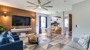 Una sala de estar moderna con un televisor grande, un panel de pared de madera, un sofá azul y una zona de comedor con una mesa y sillas.