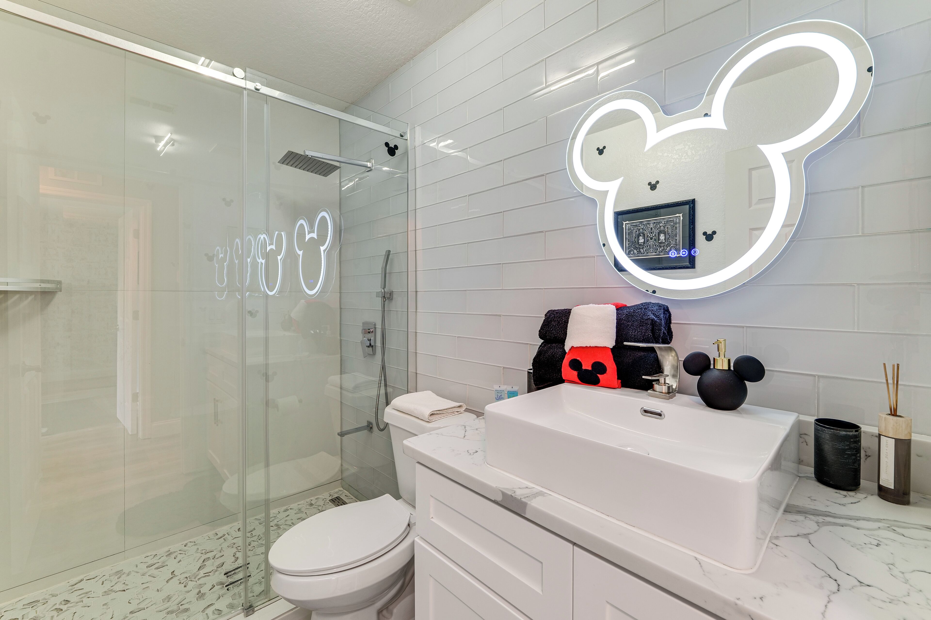 Ein Badezimmer mit Duschkabine aus Glas, Toilette, Waschbecken mit Spiegel und einer Lampenleuchte im Mickey-Mouse-Design.
