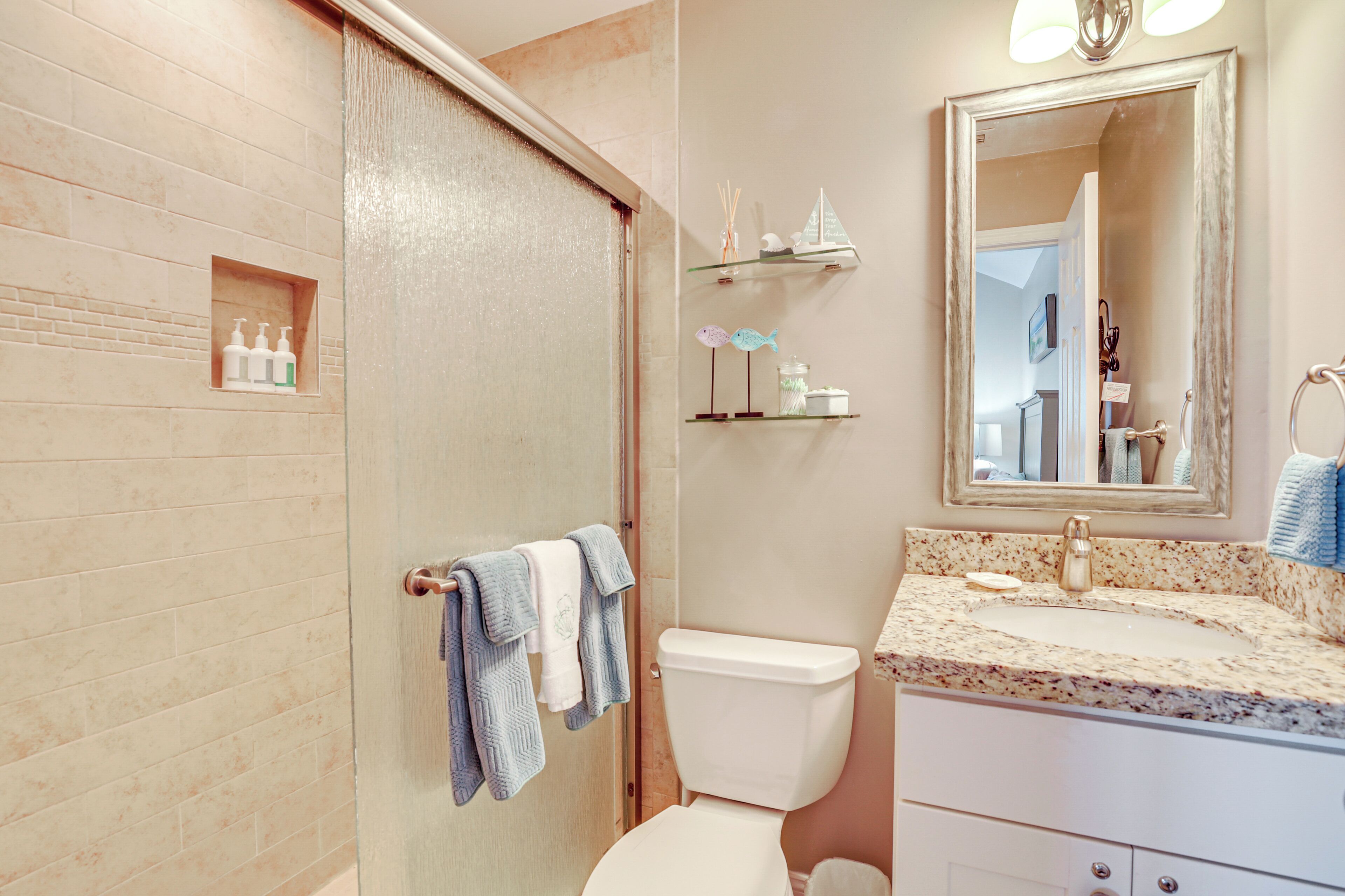 A bathroom with a shower, toilet, sink, and mirror. There are toiletries on a shelf and towels hanging on a rack.