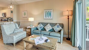 Un salon avec un canapé, un fauteuil et une table basse, un coin repas avec une table et des chaises, et une cuisine à l’arrière-plan.