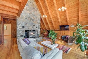 A cozy living room with a stone fireplace, wooden beams, a large sofa, and a television. - 1 Mi to Grafton Lakes St Prk: A-frame in the Woods (Cropseyville)