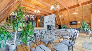 A spacious loft with a high ceiling, wooden beams, a stone fireplace, a TV, and a dining area with chairs and tables.