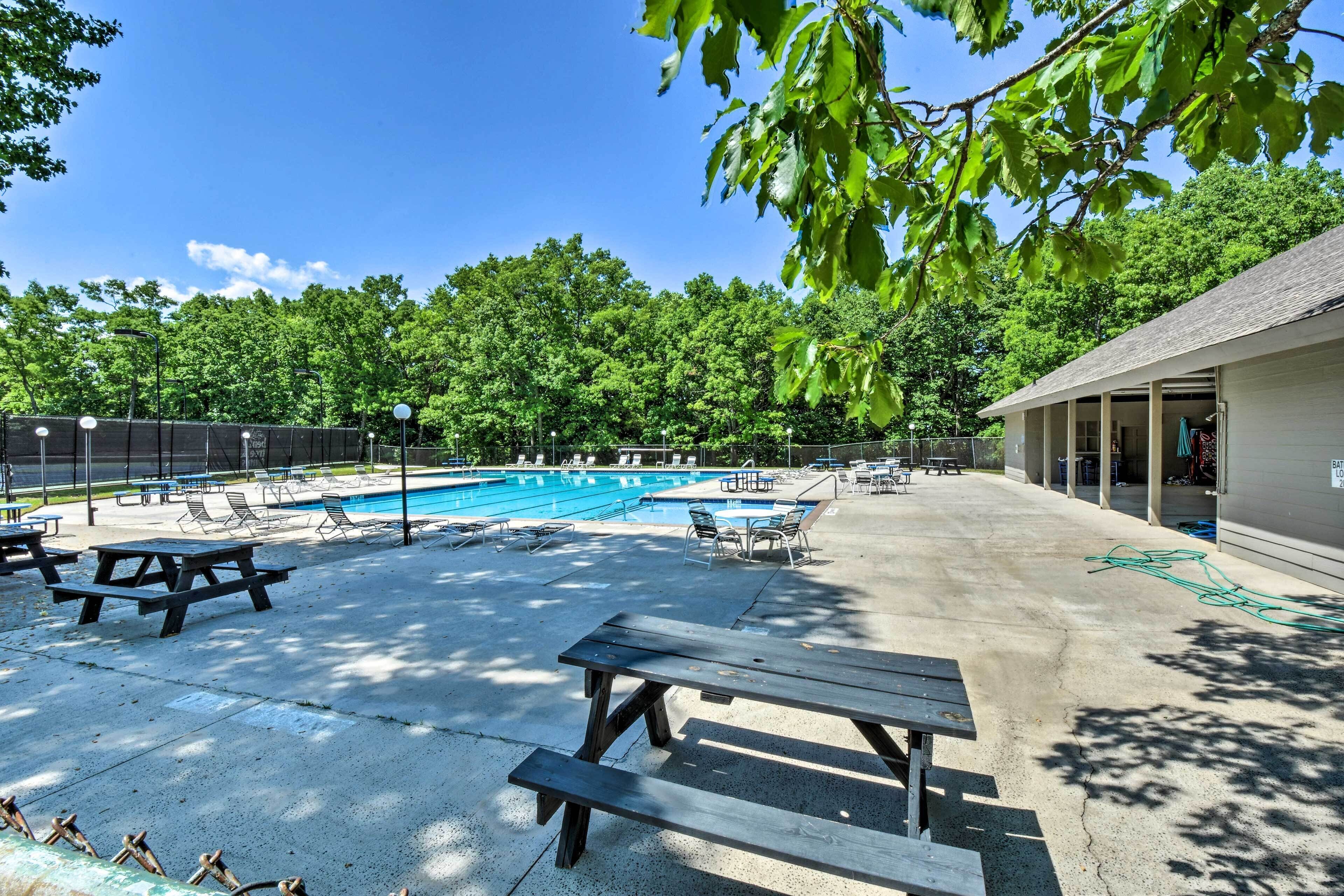 A pool area with lounge chairs and picnic tables, surrounded by trees and a building with a covered patio.