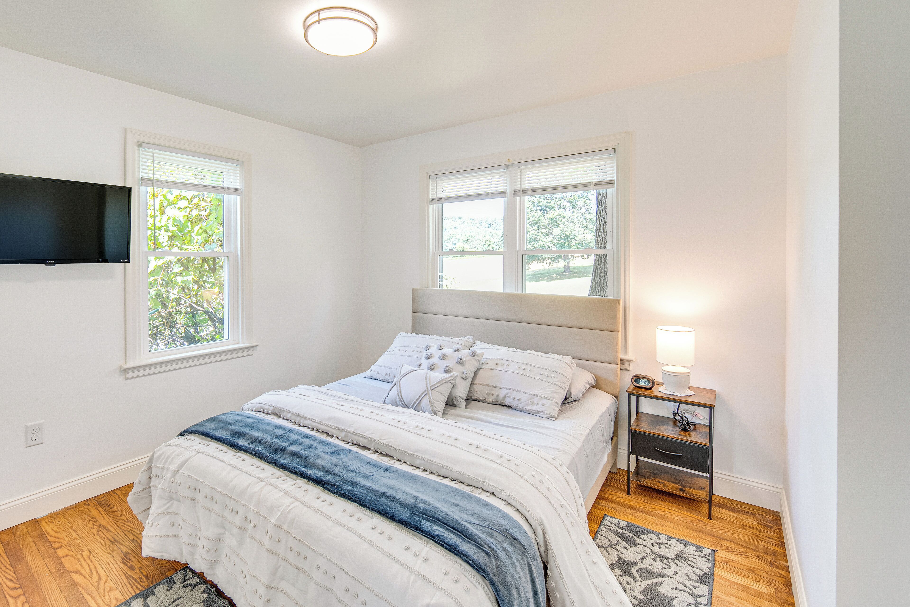 A bedroom with a bed, two windows, a nightstand, and a TV mounted on the wall.