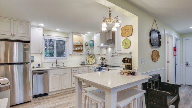 A modern kitchen with white cabinetry, a stainless steel refrigerator, a built-in oven, and a central island with bar stools. - Hot Tub & Fenced Yard: Cozy Cottage Grove Home (Cottage Grove)