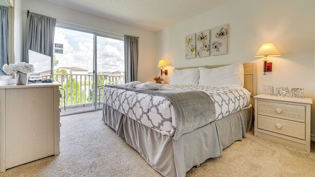 A bedroom with a bed, bedside tables, a dresser, a balcony with curtains, and artwork on the wall. - Townhouse in Lake Berkley Resort! 5 Mi to Old Town (Kissimmee)