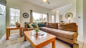 A living room with a brown leather sofa, two wooden coffee tables, a ceiling fan, and a large window with curtains. - Townhouse in Lake Berkley Resort! 5 Mi to Old Town (Kissimmee)