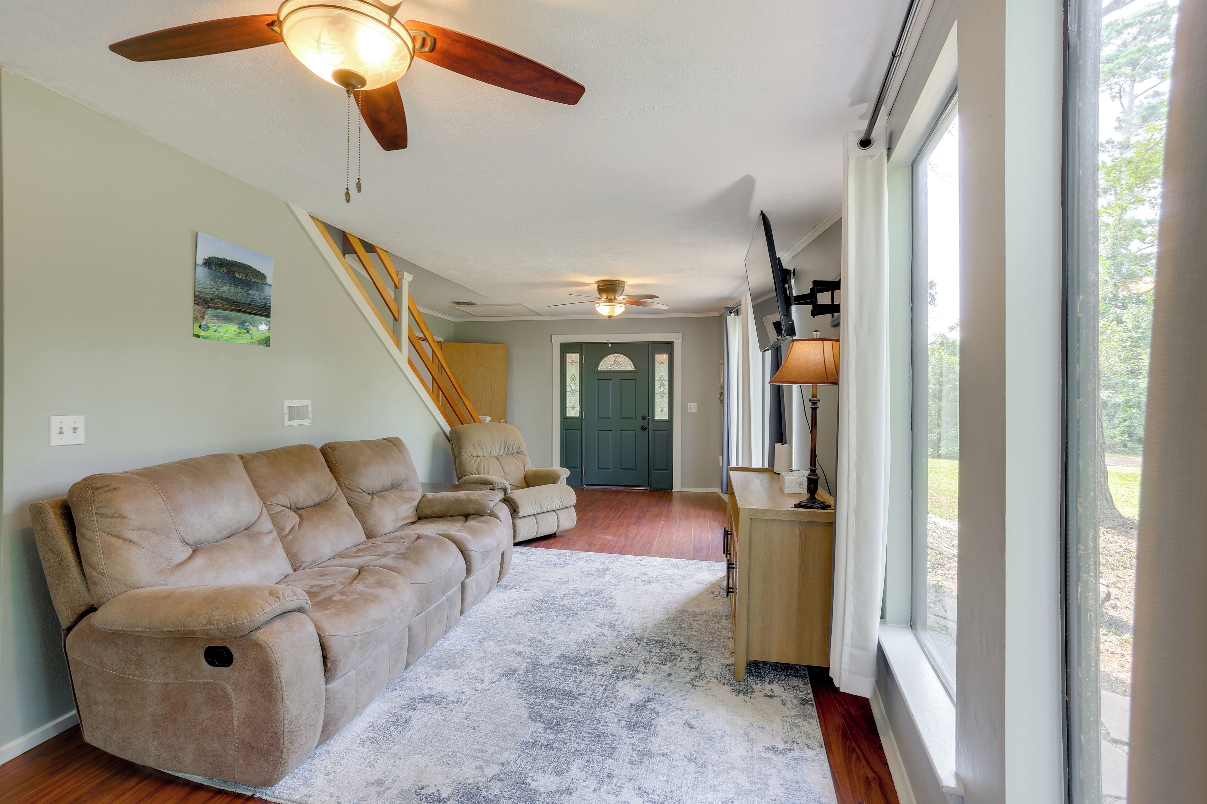 A living room with a beige sectional sofa, a ceiling fan, a wooden desk with a lamp, and a view of the outdoors through large windows.