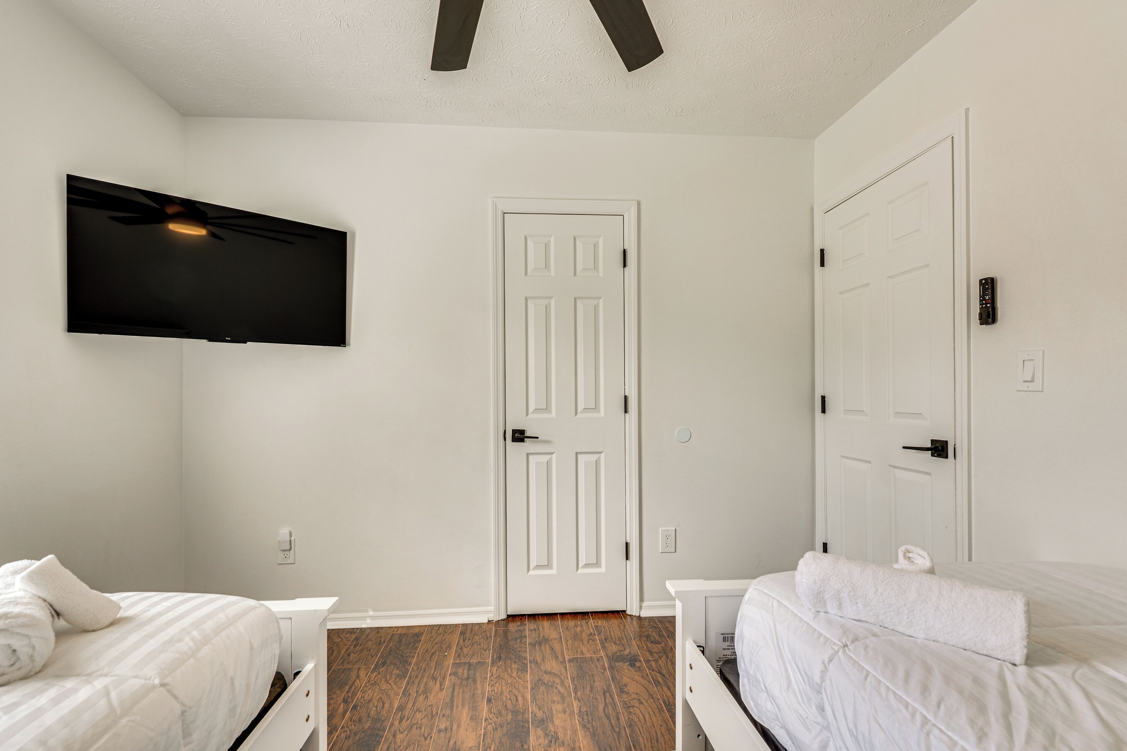 A bedroom with two beds, a ceiling fan, a mounted TV, and a white door.