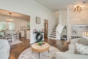 Un salon moderne avec une table basse ronde, un coin repas avec une table et des chaises blanches, et une cuisine avec des meubles blancs et des appareils en acier inoxydable.