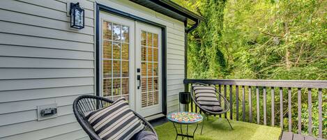A patio with two wicker chairs and a small round table, a white building with a door, and a wooden deck.
