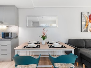 Un comedor moderno con una mesa de madera dispuesta para dos, una cocina al fondo y un sofá gris a la derecha.