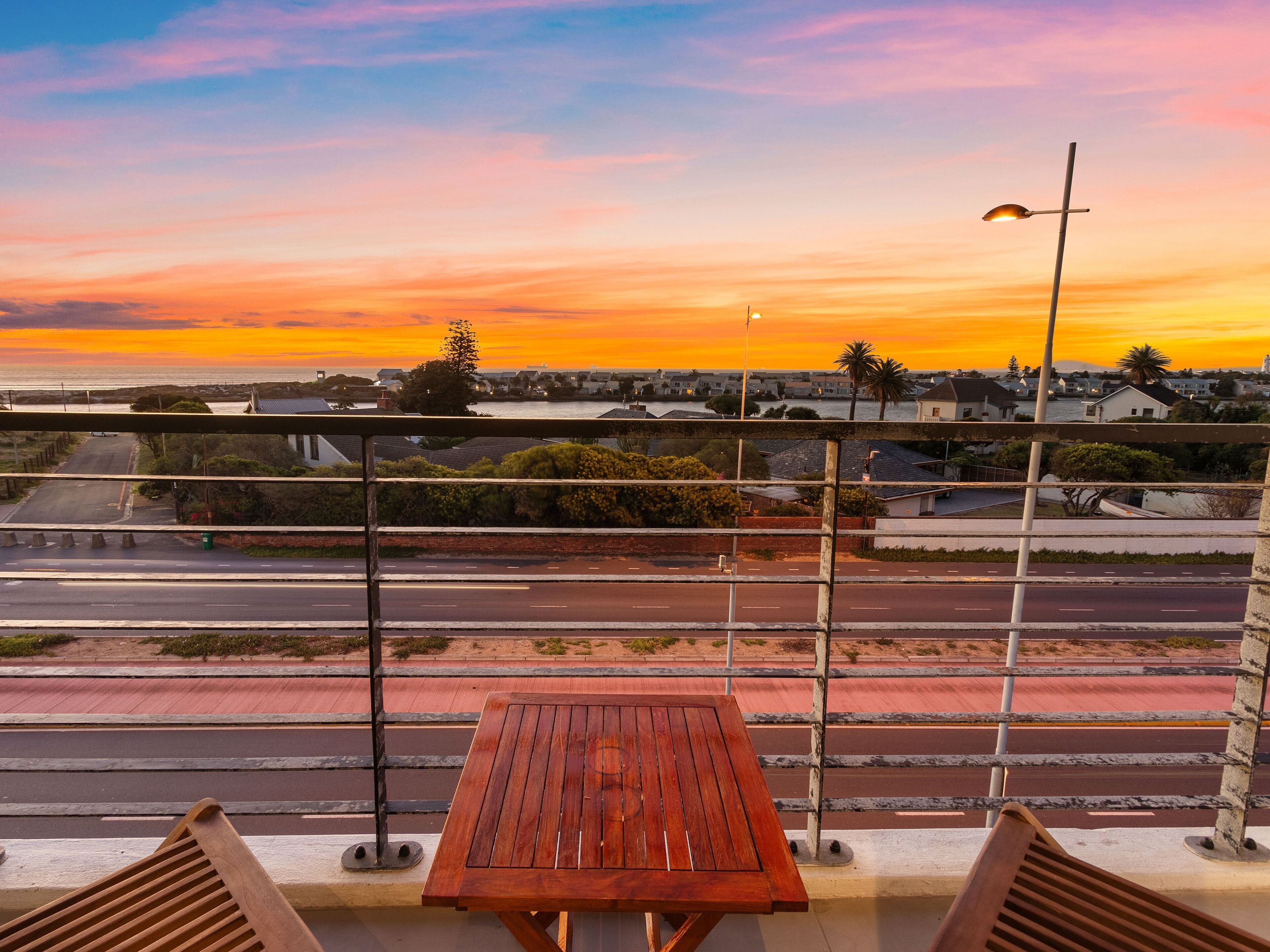 Un balcon avec une table en bois et une rambarde en métal, donnant sur une rue et offrant une vue sur le coucher du soleil.