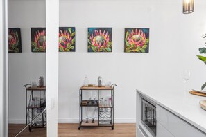 A kitchen with a white countertop, a wooden cart with glassware, and three paintings of flowers on the wall.