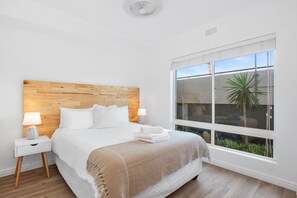A bedroom with a wooden headboard, a large window with a view of greenery, and a neatly made bed with a white comforter and a beige throw blanket. - Leisure Bay 117 (Cape Town)