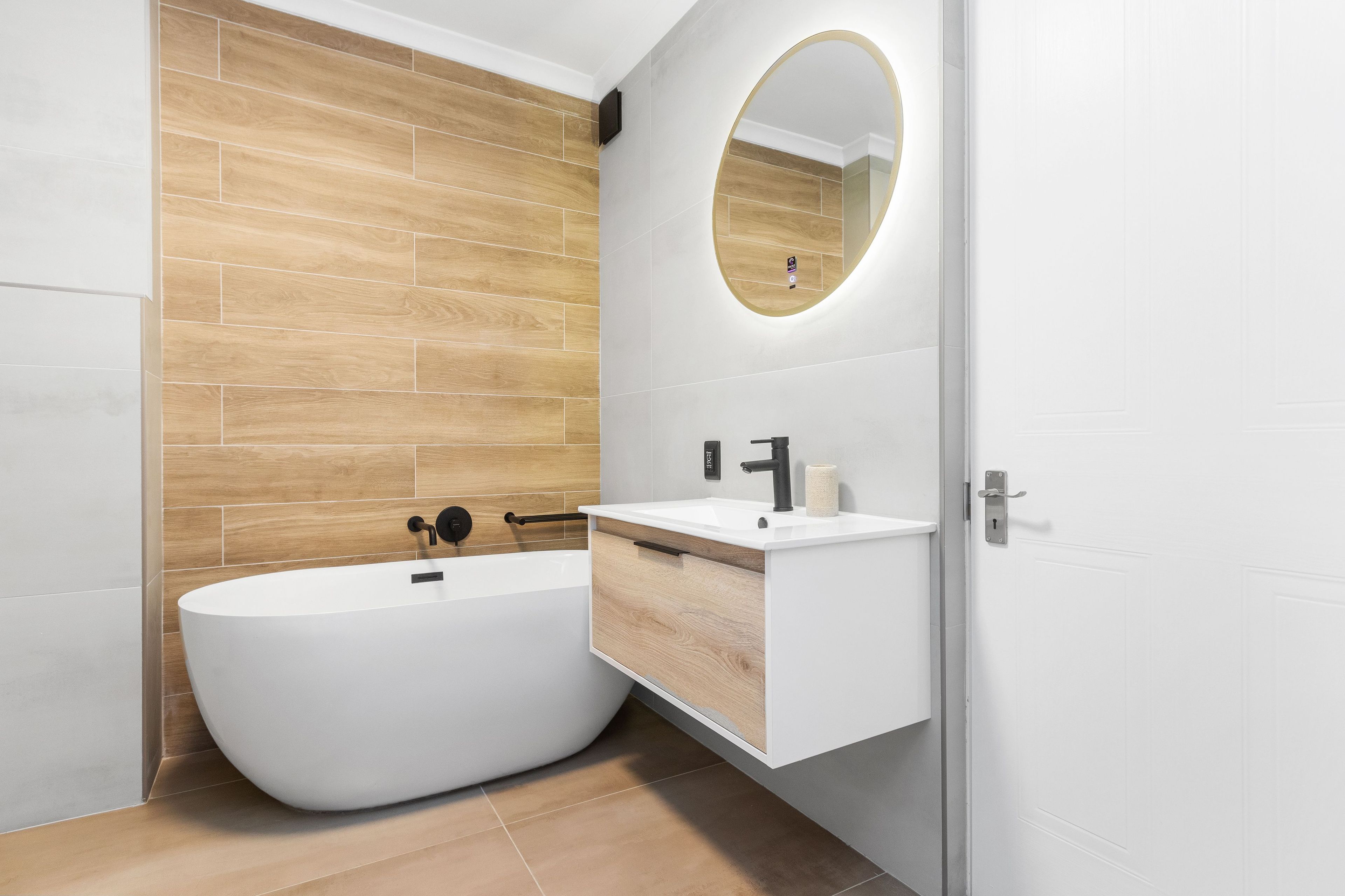 A modern bathroom with a freestanding bathtub, a wooden wall, a white vanity with a mirror, and a door.