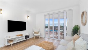 Un salón moderno con un televisor de pantalla plana, un sillón blanco, un sofá con cojines, suelo de madera y un ventanal corredero que da a un área exterior.