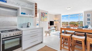 A modern kitchen with a built-in stove, white cabinetry, and a dining area with wooden stools. - Neptune Isle 9 (Cape Town)