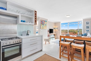 A modern kitchen with a built-in stove, white cabinetry, and a dining area with wooden stools. - Neptune Isle 9 (Cape Town)
