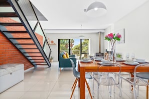 A modern dining area with a wooden table set for four, clear chairs, and a staircase with a red brick wall. - Manhattan Wharfside 301 (Cape Town)