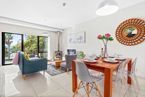 A modern living room with a dining area, large windows, and a view of greenery outside. - Manhattan Wharfside 301 (Cape Town)
