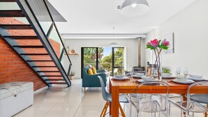 A modern dining area with a wooden table set for four, clear chairs, and a staircase with a red brick wall. - Manhattan Wharfside 301 (Cape Town)