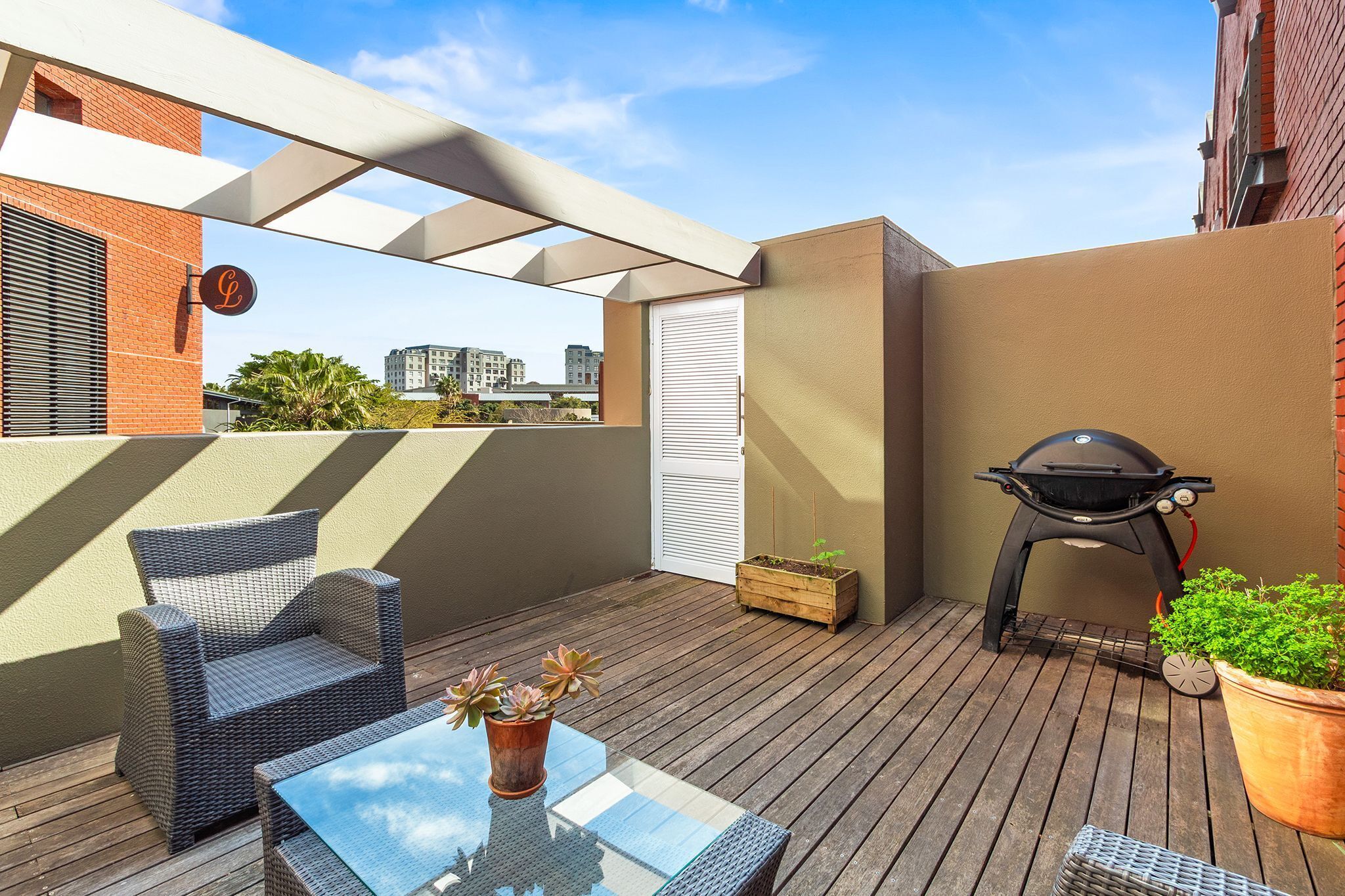 A rooftop patio with a wicker chair, a glass table, a barbecue grill, and potted plants.
