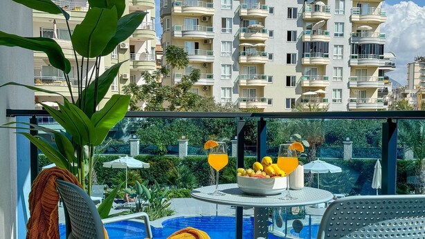 Un balcón con piscina, sillas, una mesa con fruta y bebidas, y vista a un edificio de varios pisos.