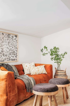 Een woonkamer met een rode bank, twee grijze poefjes, een geweven mand en een potplant.
