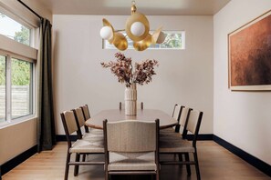A dining room with a round table, six chairs, a vase with dried flowers, a ceiling light fixture, and a painting on the wall.