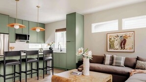 A modern living room with a sofa, coffee table, and bar stools. There is a kitchen area with green cabinets and a microwave. A painting is on the wall, and there is a window with blinds.