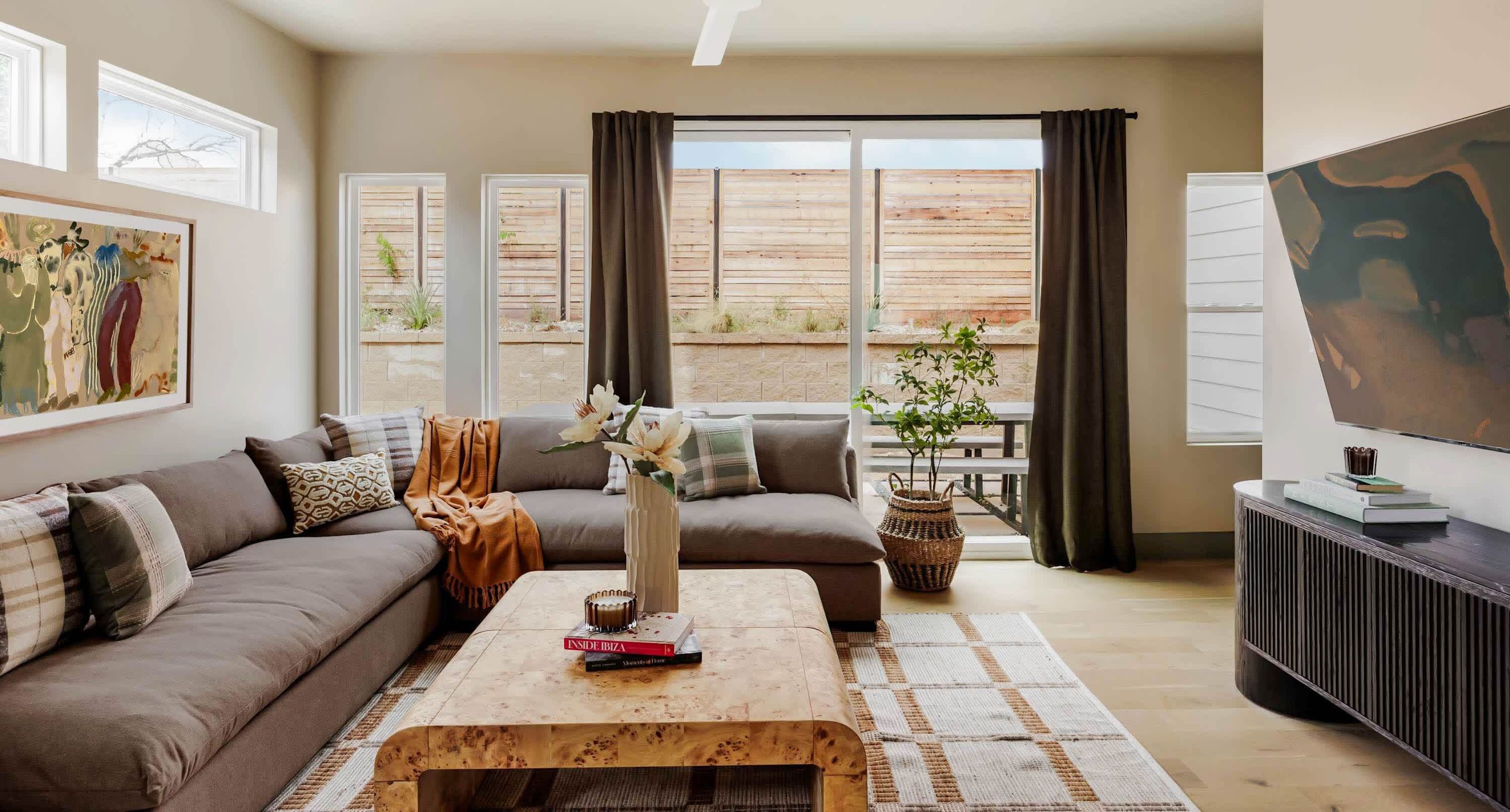 A living room with a sofa, coffee table, and a TV. There are windows with curtains, a painting on the wall, and a plant on the windowsill.