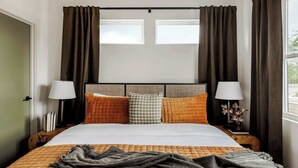 A neatly made bed with a white comforter and a gray throw blanket. There are two nightstands with lamps and a small table with a decorative item. The room has a window with curtains and a view of the outdoors.