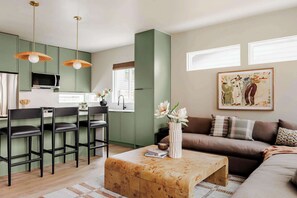 Een moderne woonkamer met een bank, salontafel en barstoelen. Er is een keukenhoek met groene kastjes en een magnetron. Een schilderij hangt aan de muur, en er is een raam met jaloezieën.