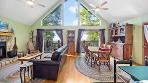 A spacious living room with a high ceiling, wooden flooring, a fireplace, a black leather sofa, a dining table with chairs, and large windows with curtains. - Above It All Cabin - Mountain Views (Murphy)