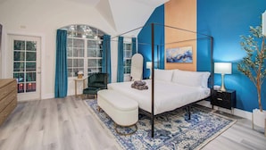 A modern bedroom with a four-poster bed, a sitting area with a chair and ottoman, a small table, and a nightstand with a lamp. - 5BD/3BA Stylish Home w Game Room, Backyard, Pool (Spring)