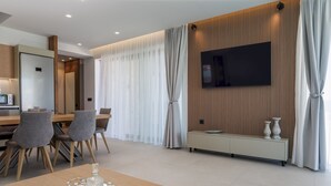 A modern hotel room with a wooden accent wall, a flat-screen TV, a dining area with a wooden table and chairs, and a seating area with a bench. - Divel Luxury Villas - Villa Ervin by Estia (Hersonissos)