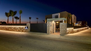 A modern building with a stone facade, a well-lit entrance, and palm trees in the background. - Divel Luxury Villas - Villa Ervin by Estia (Hersonissos)