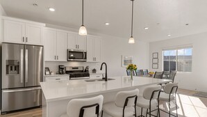 Una cocina moderna con armarios blancos, electrodomésticos de acero inoxidable, una isla central con taburetes altos, iluminación colgante y un amplio ventanal que proporciona luz natural.