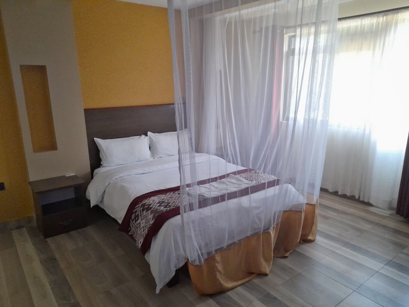 A hotel room with a bed covered by a white canopy, a wooden headboard, and a patterned throw blanket.