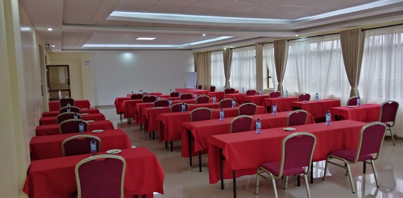 Un salón de conferencias con filas de mesas y sillas, cada una con mantel rojo y botellas de agua.