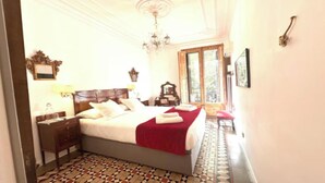 A bedroom with a large bed, a wooden nightstand, a red throw blanket, and a chandelier.