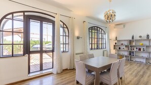 A spacious room with a large dining table, white chairs, and a modern chandelier. There are large arched windows with curtains, a wooden floor, and a bookshelf with decorative items.