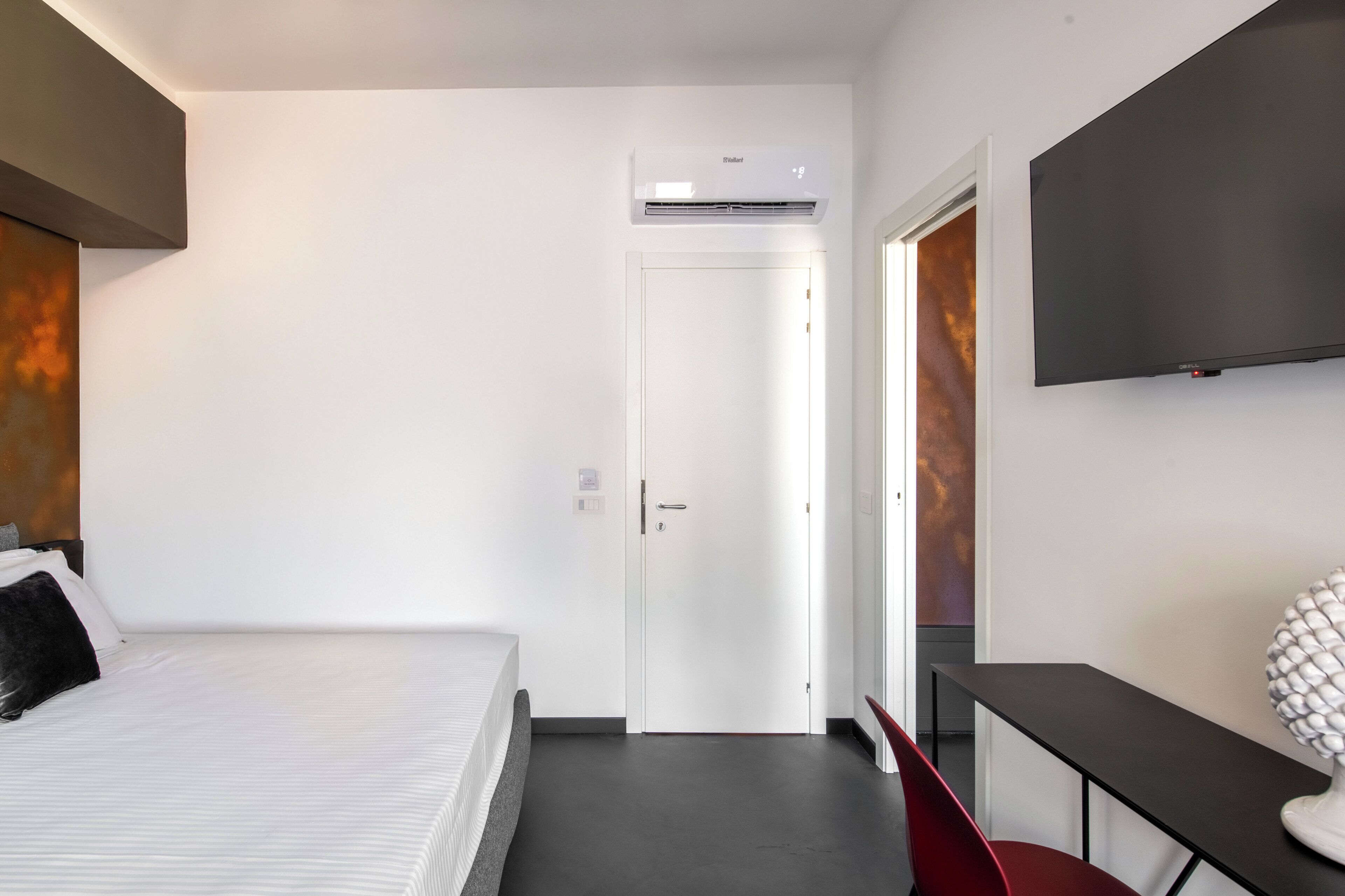 A hotel room with a bed, a desk, a chair, a TV, and an air conditioning unit.