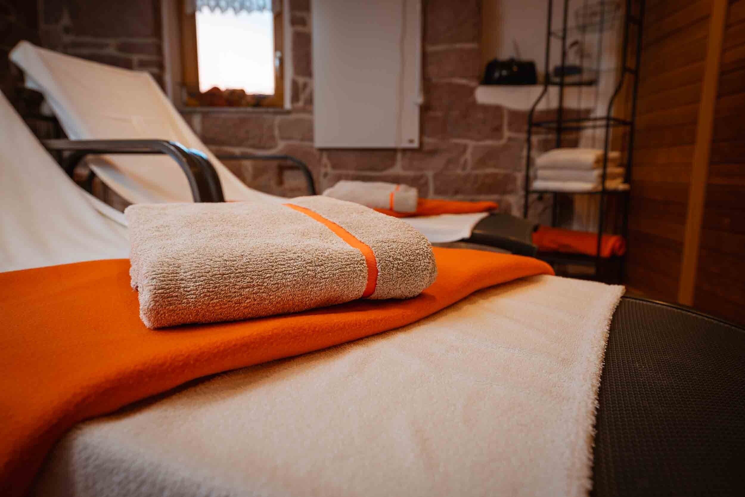 Ein Bett mit einem weißen Handtuch und einer orangefarbenen Decke, ein Metallregal mit Handtüchern und eine Backsteinwand.